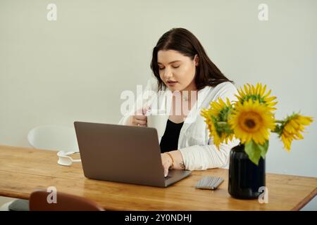 A plus size woman enjoys a warm drink while working on her laptop at a sunlit table. Stock Photo