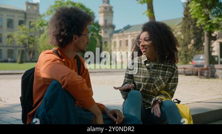 African American ethnic couple students people pupils classmates woman girl lady female man male guy sit outdoors in city talking friendly communicate Stock Photo