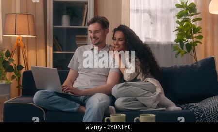 Multiracial couple shopping computer together choose e-commerce buy goods using laptop discuss at home boyfriend husband male Caucasian man guy Stock Photo