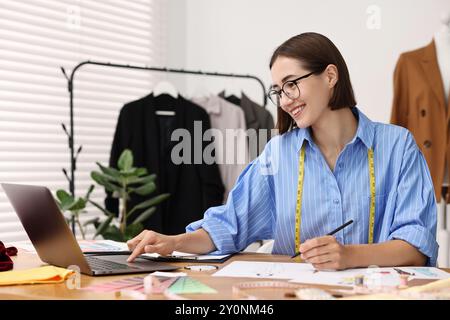 Fashion designer using laptop while drawing sketch of beautiful outfit at wooden table in workshop Stock Photo