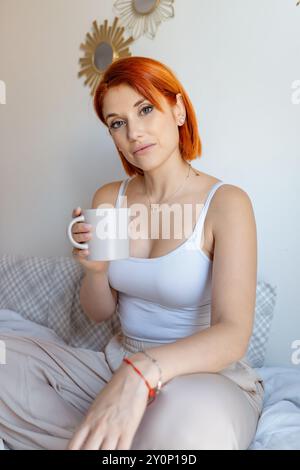 Red-haired woman sits on a bed, holding a coffee cup and looking calmly at the camera. She is dressed in a white tank top and beige pants, Stock Photo