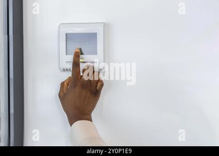 African American businesswoman uses modern digital thermostat to manage office temperature. Image depicts hand interacting with sleek device on wall, showcasing smart technology in workplace Stock Photo