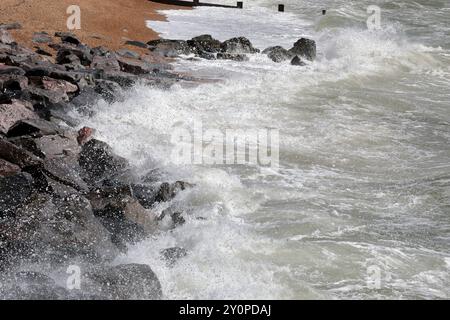 Waves breaking against the grey rocks during windy conditions at the beach, sand and groynes in the background Stock Photo
