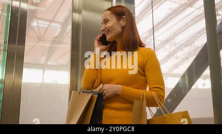 Happy laughing young Caucasian ginger woman female customer girl shopper consumer riding modern glass elevator carry shopping bags friendly talk Stock Photo