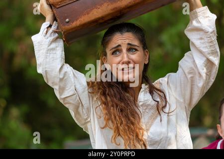 Actress in a white costume grimaces while lifting a heavy, vintage suitcase over her head during a theater performance Stock Photo