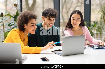 Happy international students friends sitting at desk in coworking space and using laptop, making project together Stock Photo