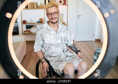 Smiling man with blond hair sitting in wheelchair in room with bookshelves and gadgets visible in background, wearing glasses and casual attire Stock Photo