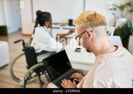 Two teammates working on laptops, collaborating in modern office setting, one using wheelchair, creating inclusive and productive atmosphere Stock Photo