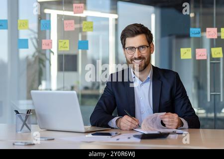 Professional businessman in office reviewing documents, using laptop and phone, surrounded by charts and sticky notes. Stock Photo