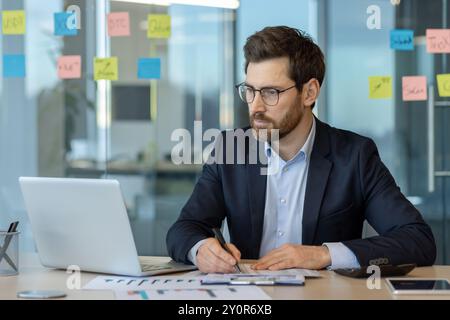 Businessman concentrating on work at desk with laptop, taking notes in modern office environment. Stock Photo