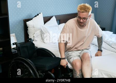Young man with light hair sitting beside wheelchair on bed with thoughtful expression Photographs on nightstand and patterned wall adding to cozy atmosphere Stock Photo