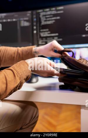 Programmer packing backpack at end of day after securing company servers from virus attacks, close up. IT remote employee finishing job shift, leaving office after building firewalls protecting data Stock Photo