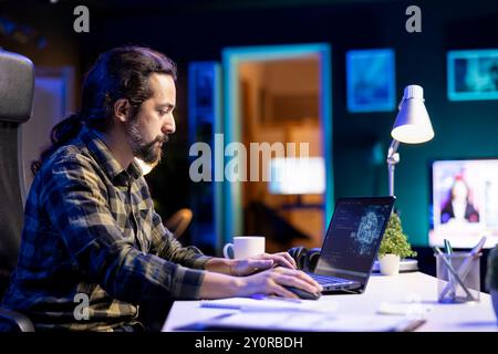 Young man managing a complex system using machine learning on his portable computer. Focused male programmer sitting and typing in code for creating neural network database on laptop. Stock Photo