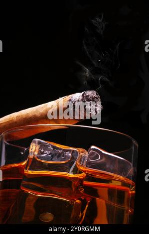 A Lit Cigar resting on Glass of Whiskey and Ice cubes with wisp of smoke and black background, low angle Stock Photo