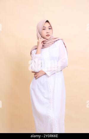 Asian woman wearing beautiful elegant muslim dress poses arms crossed holding left cheek isolated on cream background - beauty, model, studio and fash Stock Photo