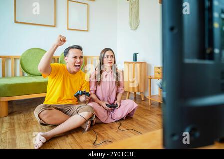 An Indonesian man and woman are playing a video game together in a living room. The man is wearing a yellow shirt and the woman is wearing a pink shir Stock Photo