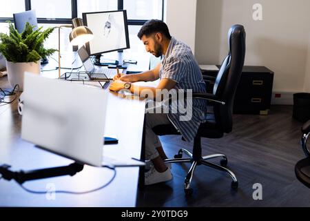 Working in modern office, Indian man writing notes at desk with computer and lamp Stock Photo