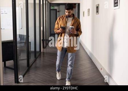 Walking in office, Indian man using smartphone and holding coffee cup Stock Photo