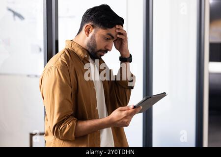 Worried Indian man holding tablet, reading important information in modern office Stock Photo