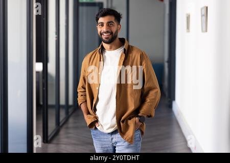 Smiling Indian man in casual attire standing in modern office hallway, hands in pockets Stock Photo