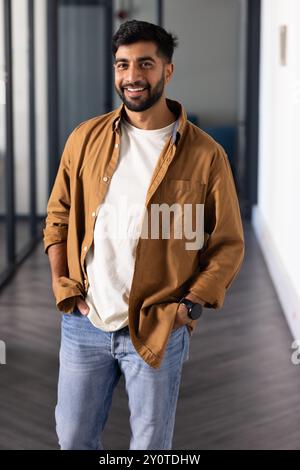 Smiling Indian man standing in office hallway with hands in pockets, wearing casual outfit Stock Photo