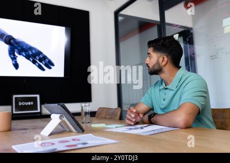In office, Indian man watching robotic hand presentation on screen, holding pencil Stock Photo