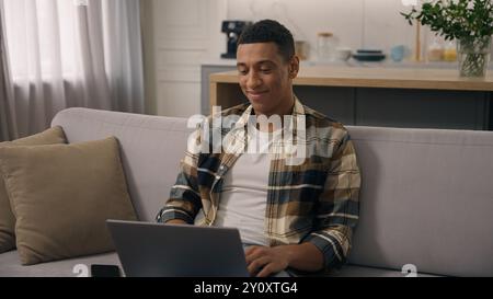 Happy smiling African American ethnic man freelancer working at home distant online work at sofa smile positive student guy studying remote with Stock Photo