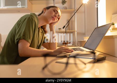 Tired young woman is sitting at her desk with a laptop and falling asleep. Fatigue due to overworking. Student falls asleep near the computer. Blurred Stock Photo