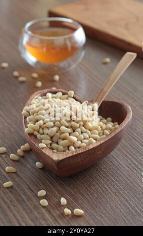 Natural pine nuts in heart shape bowl. Wooden dishes and furniture in kitchen. Fresh clear honey in glass bowl. Healthy products.  Stock Photo