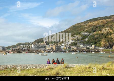 Rear view of four people sitting on the beach looking over the Mawddach estuary from Penrhyn Point, Fairbourne to Barmouth, Gwynedd, North Wales, UK. Stock Photo