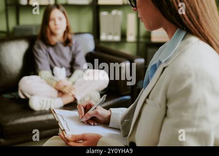 Therapist wearing suit taking notes while counseling young woman sitting on couch in office providing professional support and advice for client's men Stock Photo