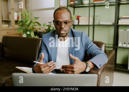Portrait of serious African American businessman working on laptop in office setting, taking notes and gesturing while participating in virtual meetin Stock Photo
