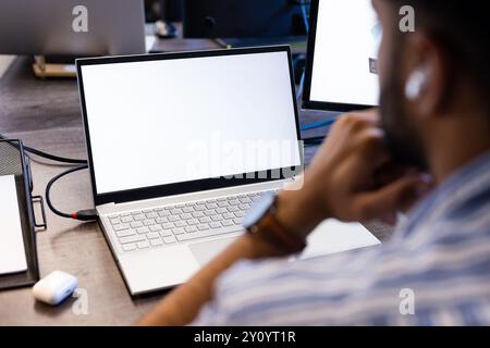 Using laptop with blank screen, Indian man thinking and wearing wireless earbuds, copy space Stock Photo