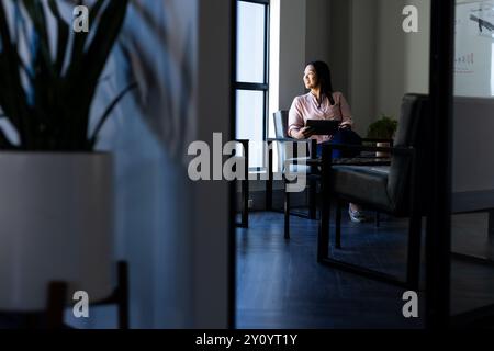 Sitting in office, woman holding tablet and looking out window, thinking Stock Photo