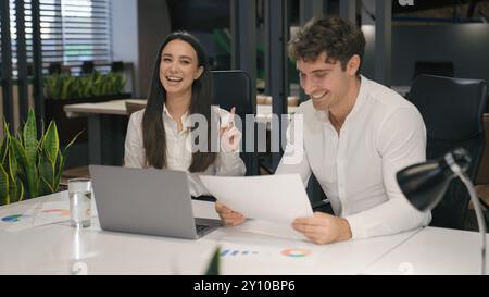 Caucasian woman and man working together with papers and laptop computer smile laughing happy fun work paperwork laugh in office business colleagues Stock Photo