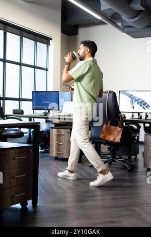 Walking in office, Indian man drinking coffee and holding tablet near desk with computer Stock Photo