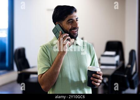 Talking on smartphone and holding coffee cup, Indian man smiling in office setting Stock Photo