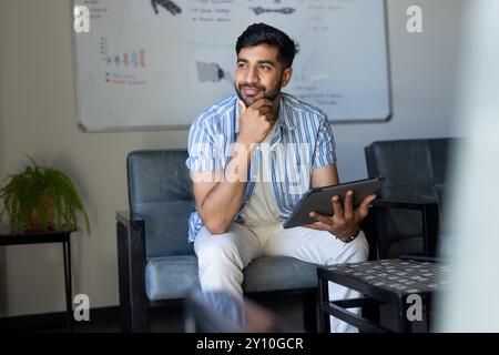 Holding tablet, Indian man sitting in office, thinking and looking away Stock Photo