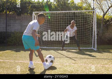 Playing soccer, boy kicking ball towards woman as goalkeeper on field Stock Photo