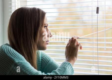 Young Woman peering through blinds sitting indoors on a sunny day, lost in thought and reflection on her surroundings Stock Photo