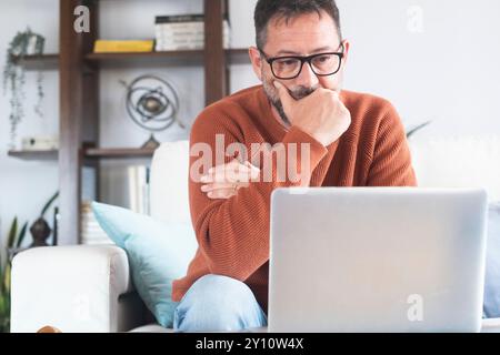 Sad and frustrated man feeling tired and worried about a problem sitting on sofa at home with laptop, stressed 50 year old male having problems reading bad news online, email notification about debt or negative message Stock Photo