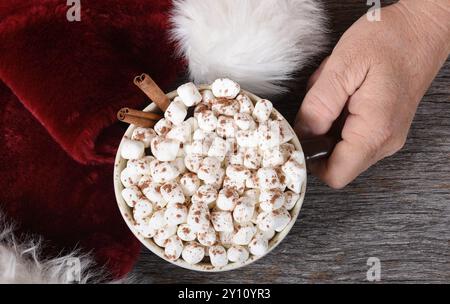 Santa Claus hand with a mug of hot chocolate next ot his traditional hat on a rustic wood table. Stock Photo