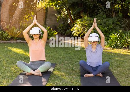 Practicing yoga outdoors, grandmother and granddaughter using vr headsets for virtual meditation Stock Photo