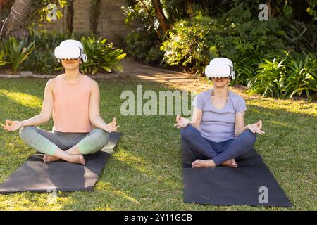 Practicing yoga on mats, asian grandmother and granddaughter wearing VR headsets in outdoor garden Stock Photo