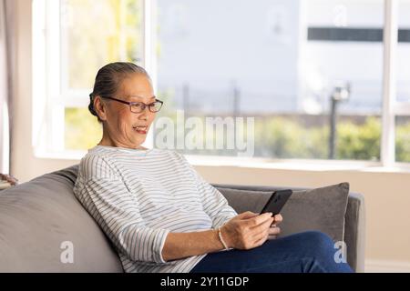 Sitting on couch, elderly Asian woman using smartphone and smiling at screen Stock Photo