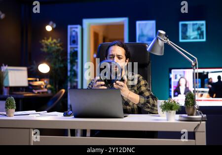 Male university student taking his wireless headset from table for music while using his laptop. Bearded man holding his headphones while seated in front of his portable computer. Stock Photo