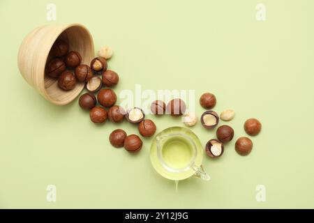Jug of macadamia oil and overturned bowl with nuts on green background Stock Photo