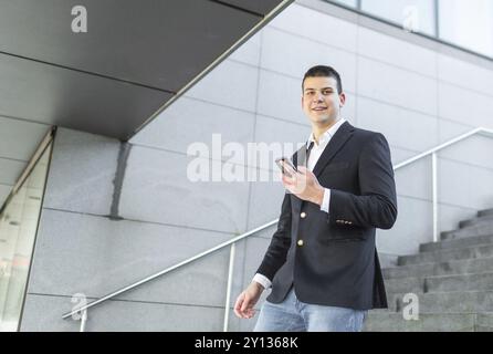 Young and Casual Businessman Using Smartphone while Walking Outside of Office Building Stock Photo