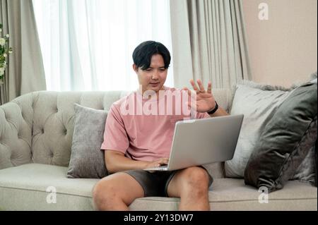 An Asian man in casual clothes is working remotely from home, having an online meeting via laptop with his colleagues while sitting on a couch in the Stock Photo
