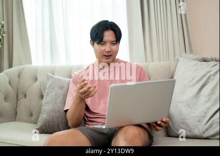 An Asian man in casual clothes is working remotely from home, having an online meeting via laptop with his colleagues while sitting on a couch in the Stock Photo